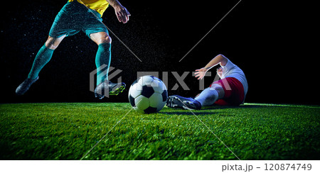 Soccer player in green kicks ball fiercely, while on green football field while other team's defender tries to stop him against black studio background. Soccer player in green kicks ball fiercely, while on green football field while other team's defender tries to stop him against black studio background. 120874749