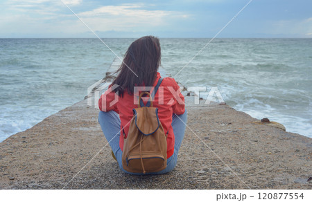 Rear view of a young woman tourist with a backpack sitting on a pier and waves hitting the pier in a storm. City of Sudak, Crimea 120877554