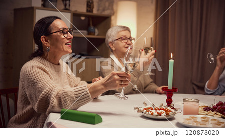 Smiling elderly women sitting table decorated with candles and plate with snacks, appetizers, drinking wine, saying toast and celebrating 120878020