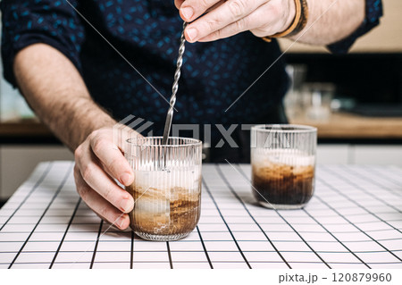 Close-up of hands stirring creamy iced coffee in ribbed glass. 2025 color Mocha Mousse Close-up of hands stirring creamy iced coffee in ribbed glass. 2025 color Mocha Mousse 120879960