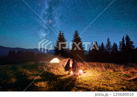 Couple hikers shares kiss by campfire, seated in chairs under magnificent starry sky with Milky Way visible above. Illuminated tent and serene mountain backdrop enhance romantic, tranquil ambiance. Couple hikers shares kiss by campfire, seated in chairs under magnificent starry sky with Milky Way visible above. Illuminated tent and serene mountain backdrop enhance romantic, tranquil ambiance. 120880010