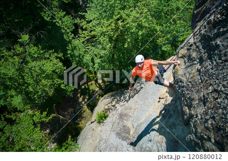 Aerial view of male rock climber ascending rugged limestone cliff with harness and rope for safety. Sportsman climbing on vertical large boulder at Dobvush Rocks in Carpathian mountains, Ukraine. Aerial view of male rock climber ascending rugged limestone cliff with harness and rope for safety. Sportsman climbing on vertical large boulder at Dobvush Rocks in Carpathian mountains, Ukraine. 120880012