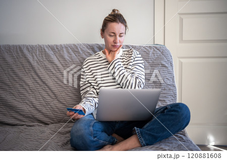 Doubtful woman on couch with credit card thoughtful using laptop pondering bills, utilities payments Doubtful woman on couch with credit card thoughtful using laptop pondering bills, utilities payments 120881608