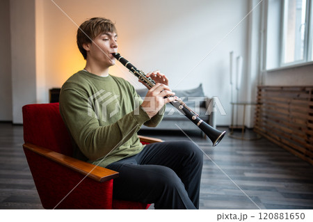 Clarinet player. Man clarinetist playing flute woodwind music instrument sitting on armchair at home Clarinet player. Man clarinetist playing flute woodwind music instrument sitting on armchair at home 120881650