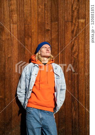 Relaxed skinny long-haired male with eyes closed leaning on wooden wall outdoors in bright sunny day 120881851