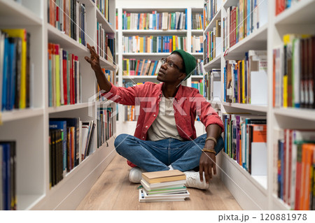 Young African American guy sits on floor in library between bookshelves, student searching for book 120881978