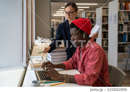 Woman teacher pointing at laptop screen while explaining something to African student guy in library 120882009