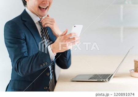 スマホを見て喜ぶ若い男性ビジネスマン（ネット詐欺・闇バイト・転売・スマホ詐欺・高額当選詐欺） 120891074