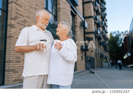 Loving retired couple in casual clothes enjoying coffee standing on city street, smiling and talking, savoring togetherness in urban landscape on sunny day, on blurred background of building exterior. Loving retired couple in casual clothes enjoying coffee standing on city street, smiling and talking, savoring togetherness in urban landscape on sunny day, on blurred background of building exterior. 120892287