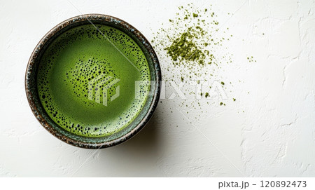 A close-up view of a rustic bowl filled with vibrant green matcha tea, accompanied by a sprinkle of matcha powder on a light textured surface, ideal for healthy beverage promotion. 120892473