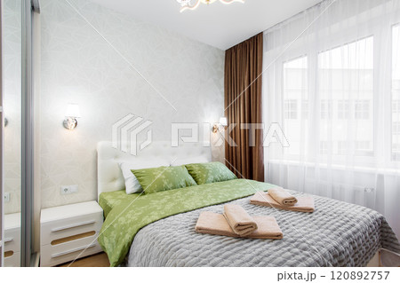 A well-appointed small modern bedroom with a cozy design, featuring a queen-sized bed, bedside tables. A large window with curtains allowing soft daylight. A well-appointed small modern bedroom with a cozy design, featuring a queen-sized bed, bedside tables. A large window with curtains allowing soft daylight. 120892757