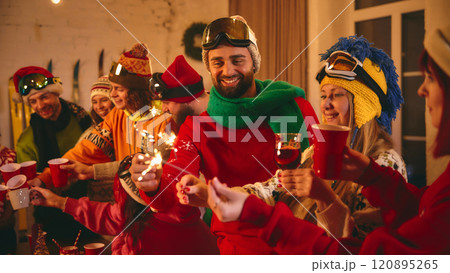 Joyful holiday celebration as friends in vibrant ski gear toast and laugh around rustic table lit with candles and sparkles, fireworks. 120895265