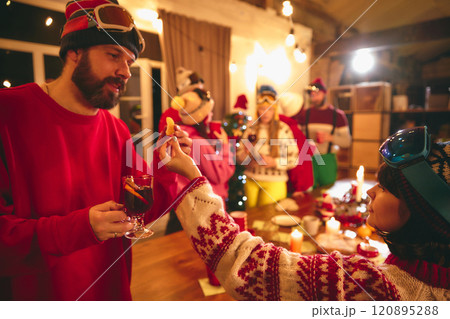 Cheerful group in winter sweaters gathers around rustic table laughing in festive mood. Couple talking and drinks hot, warming drinks. 120895288