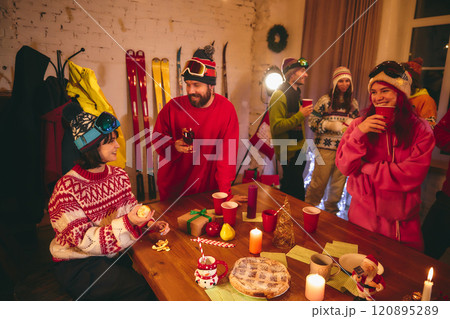 Festive spirit fills room as friends in ski outfits share drinks and laughter around holiday-themed table with candles, gifts, and seasonal treats. 120895289