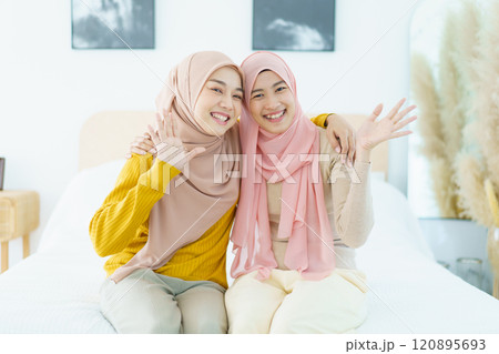 Portrait of Asian beautiful happy Muslim women relaxing in their house together. Cheerful Muslim female sibling relaxing - sitting on the bed then waving a hands and smile to camera. 120895693