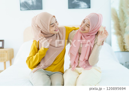 Portrait of Asian beautiful happy Muslim women relaxing in their house together. Cheerful Muslim female sibling relaxing - sitting on the bed then waving a hands and smile to camera. 120895694