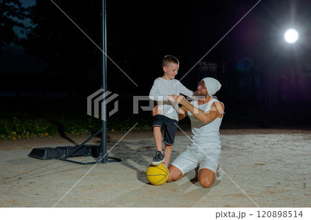 Happy family dad and son looking at camera and smiling while sitting on basketball court outdoor. Boy with handsome father after game on playground, sport concept Happy family dad and son looking at camera and smiling while sitting on basketball court outdoor. Boy with handsome father after game on playground, sport concept 120898514