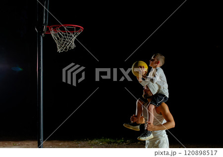 A father joyfully lifts his son for a basketball shot under the night sky at a local court A father joyfully lifts his son for a basketball shot under the night sky at a local court 120898517