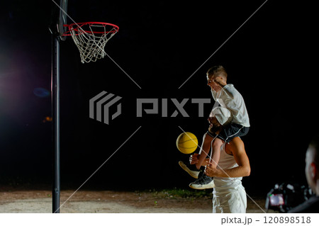 A father joyfully lifts his son for a basketball shot under the night sky at a local court 120898518