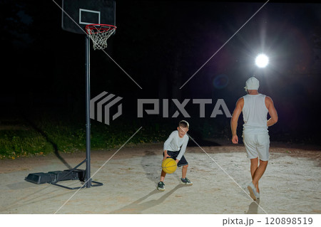 A nighttime basketball game between a child and an adult on an outdoor court under bright lights A nighttime basketball game between a child and an adult on an outdoor court under bright lights 120898519