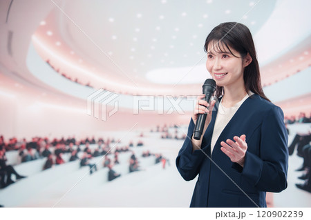 ホールで司会・プレゼンをするスーツを着た若い女性 120902239