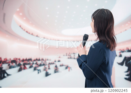 ホールで司会・プレゼンをするスーツを着た若い女性 120902245