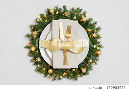 Beautiful Christmas table setting with gold cutlery and decorations on white background. Plate in a New Year wreath with a golden bow and garland lights. Beautiful Christmas table setting with gold cutlery and decorations on white background. Plate in a New Year wreath with a golden bow and garland lights. 120903453
