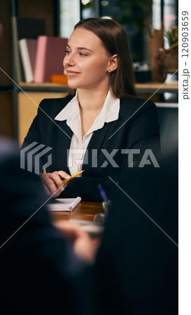 Confident young businesswoman in formal attire attentively listening during professional meeting, holding pen and notebook. Confident young businesswoman in formal attire attentively listening during professional meeting, holding pen and notebook. 120903659
