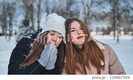 Two schoolgirls standing in a snowy park, warming hands during winter snowfall, bundled up in warm clothing. Happy and cheerful, showcasing friendship in the cold 120907309