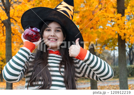 Smiling girl in witch hat holding candy jar with autumn leaves background Smiling girl in witch hat holding candy jar with autumn leaves background 120912219
