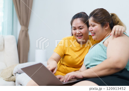 Happy Asian women friend relaxing in living room and drink a cup of tea together. Happy - delighted two Asian oversized women talking and watching a movie on laptop or notebook computer. Happy Asian women friend relaxing in living room and drink a cup of tea together. Happy - delighted two Asian oversized women talking and watching a movie on laptop or notebook computer. 120913452