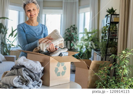 Gray haired mature smiling woman pack clothes for recycling to cardboard box at home 120914115