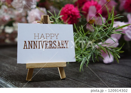 Happy Anniversary text write on paper card with wooden easel and flowers decoration on wooden background 120914302