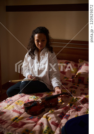 Woman sitting on bed admiring violin, basked in soft evening light 120929872
