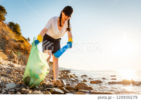 Bottom view of young Caucasian woman in rubber gloves collects garbage on pebble wild beach. Volunteer puts garbage in plastic bag. Concept of save Earth and environment disaster 120930279