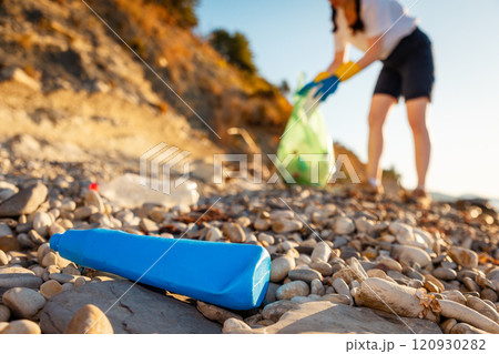 Close up of plastic blue bottle on coast. Defocused volunteer collects garbage on pebble beach in background. Concept of ocean pollution and ecological disaster 120930282