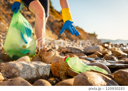 Close up of volunteer hand in rubber gloves pickups plastic bottle on pebble wild beach. Concept of ocean's pollution and clean up of coast 120930283