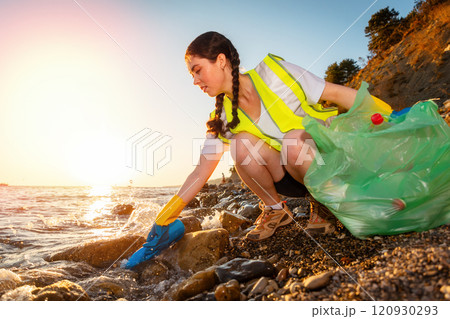 Low angle view of young Caucasian woman volunteer in rubber gloves collect garbage squatting on coast. Concept of Earth Day and ocean seashore cleanup 120930293