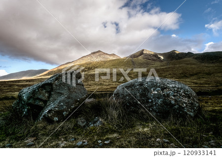 Two large, dark rocks sit in grassy foreground. Mountains rise in background under a partly cloudy sky. Sunlight illuminates mountain peaks. Two large, dark rocks sit in grassy foreground. Mountains rise in background under a partly cloudy sky. Sunlight illuminates mountain peaks. 120933141