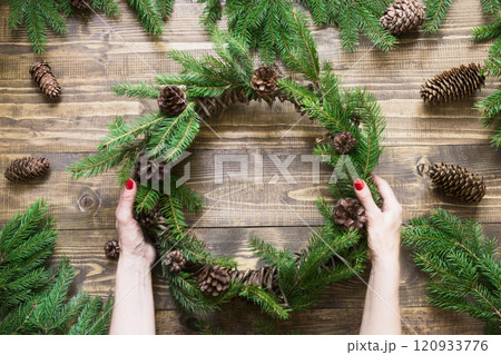 Making Christmas wreath using fresh and all natural materials. Christmas wreath with fir branches on wooden board in female hand. Flat lay. Top view. 120933776