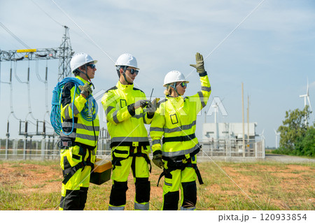 Three wind turbine or windmill workers or engineer stay in front of small electricity or power plant and some of them use mobile phone to check the data with wind turbine as background. 120933854