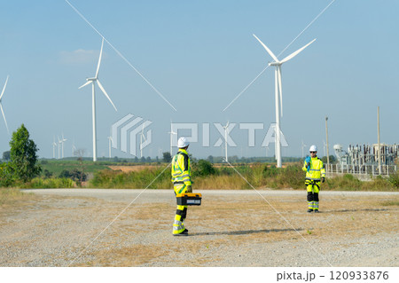 Two wind turbine or windmill workers walk and discuss together on road during work in workplace field in day time and wind turbine are in the background. 120933876