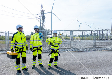Three wind turbine or windmill workers or engineers stay on road near small electricity or power plant and look to right side and wind turbine as background. 120933895