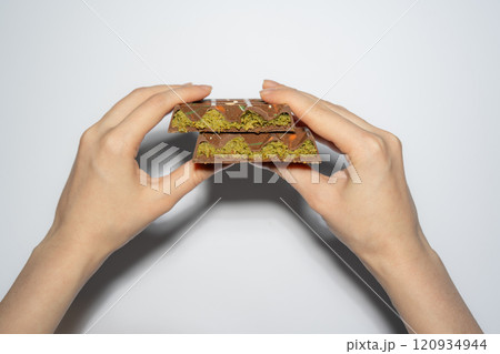 Trend Dubai chocolate bar with pistachio paste, tahini and kataifi dough in woman's hands. Confectionery and sweets. Trend Dubai chocolate bar with pistachio paste, tahini and kataifi dough in woman's hands. Confectionery and sweets. 120934944