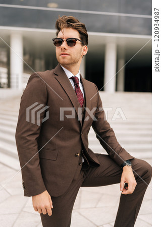 Vertical portrait of serious confident businessman in stylish suit and sunglasses posing outdoors, exuding success and professionalism, city architecture complementing modern urban look. Vertical portrait of serious confident businessman in stylish suit and sunglasses posing outdoors, exuding success and professionalism, city architecture complementing modern urban look. 120934987