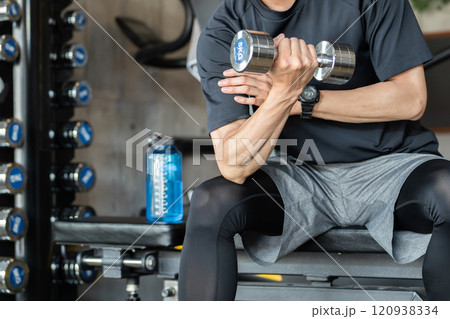 ジムのトレーニングベンチに座りながら筋トレをする若いアジア人男性（腕トレ・ダンベル・ダンベルカール） 120938334