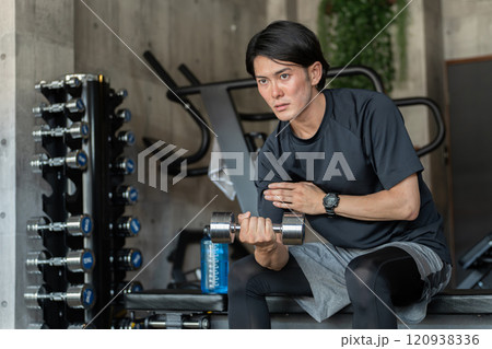 ジムのトレーニングベンチに座りながら筋トレをする若いアジア人男性(腕トレ・ダンベル・ダンベルカール) ジムのトレーニングベンチに座りながら筋トレをする若いアジア人男性(腕トレ・ダンベル・ダンベルカール) 120938336