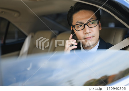 後部座席に座りスマホで話すビジネスマン 120940896