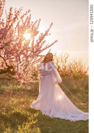 Woman blooming peach orchard. Against the backdrop of a picturesque peach orchard, a woman in a long white dress enjoys a peaceful walk in the park, surrounded by the beauty of nature. 120941880