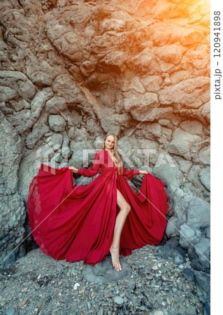 Red dress rocks woman. A blonde with flowing hair in a long flowing red dress stands near a rock of volcanic origin. Travel concept, photo session at sea Red dress rocks woman. A blonde with flowing hair in a long flowing red dress stands near a rock of volcanic origin. Travel concept, photo session at sea 120941898